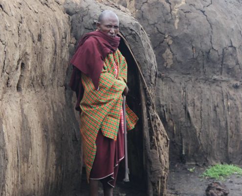 While on your 7 Days Safari Tanzania you will have the chance to encounter and learn about local people & culture like this elder Maasai standing in front of a traditional Boma made from adobe.