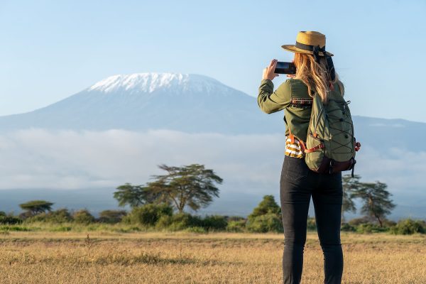 beautiful young girl In a hat stands against the backdrop of the Kilimanjaro volcano and looks away. The concept of tourism and African safari. female backpacker takes pictures of nature and animals