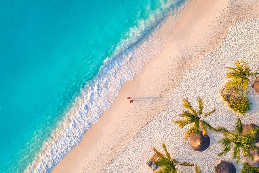 Aerial view of umbrellas, palms on the sandy beach of Indian Ocean at sunset. Summer in Zanzibar, Africa. Tropical landscape with palm trees, parasols, walking people, blue water, waves. Top view